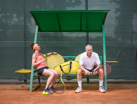 Tennis players are resting on the bench between sets. Mixed age. Sports family. Young woman and elderly man. Mixed gender match