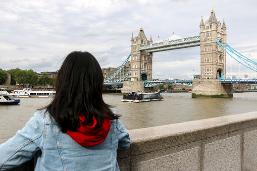 Female Tourist in London, UK, looking at one of the most iconic sights of the city, the Tower Bridge as tour boats cruise pass along the River Thames
