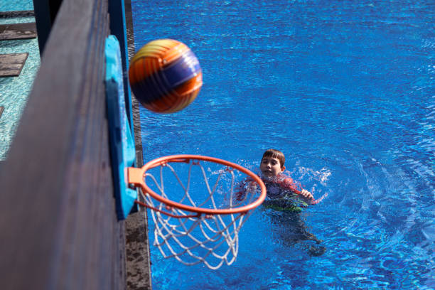 a school-age boy throws a ball into a basketball basket in a swimming pool