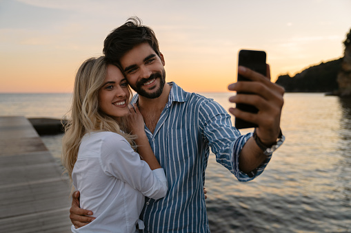 Lovely couple using mobile phone at the beach. Two attractive people enjoying an evening, walking and taking a selfie on vacation.