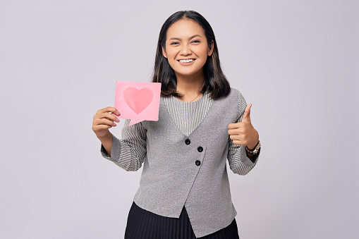 Smiling happy young Asian woman holding a heart form-like icon sign from social network media feedback and showing thumb up sign isolated on grey background