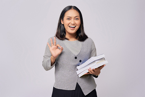 Beautiful smiling young Asian woman looking smart standing confident with holding books on hand while looking at the camera and showing okay sign isolated on grey background