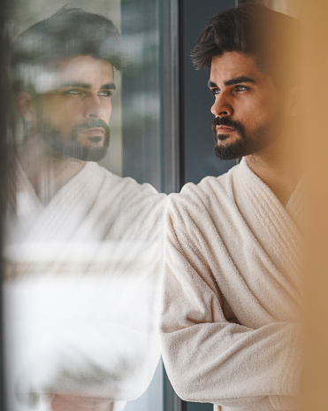 Side view close-up photo of good looking serious man with dark hair and light eyes standing next to window wearing bathrobe.