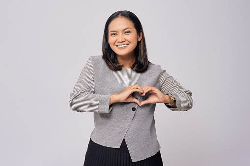 Portrait of a happy young Asian woman in formal wear showing heart gesture, making love signs with fingers and looking at the camera with smiling face isolated on grey background