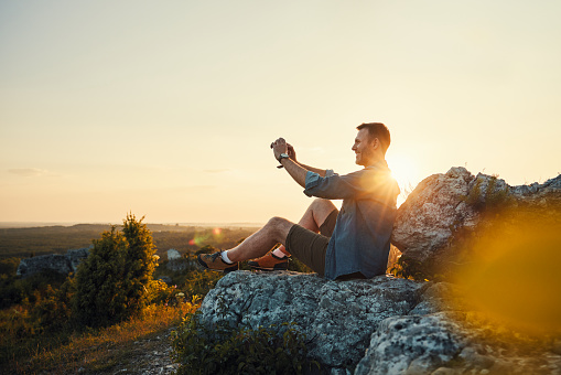 Male hiker taking photo sitting on the rock during sunset. Hiking travel concept.