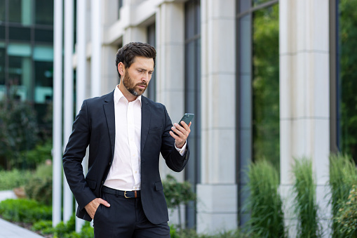 Serious thinking businessman boss holding phone, man reading online message and typing, investor outside office building, successful financier concentrated banker, using app on smartphone.