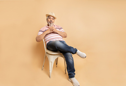 Retired senior indian man casual cloths and hat drinking soda water in plastic glass while sitting on chair relaxing isolated on beige studio background. Hot summer season, Retirement life.