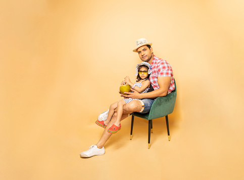 Young indian father and his little daughter drinking coconut water while sitting on chair relaxing isolated on beige studio background. Holidays and vacations, Summer season, Full length, Copy space