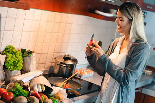 Young happy woman cooking dinner at home. Smiling blonde girl holding knife and chopping vegetable over the pan while standing next to stove.