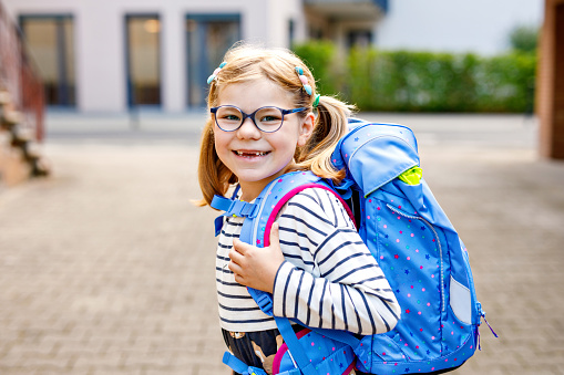 Cute little girl on her first day going to school. Healthy beautiful child walking to nursery preschool and kindergarten. Happy child with eyeglasses with backpack on the city street, outdoors