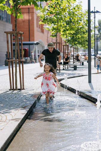 Little happy Ukrainian girl meets her father from the war during his vacation and cheerful plays with him near fountain in city. War in Ukraine. Russian military invasion in Ukraine. War and children.