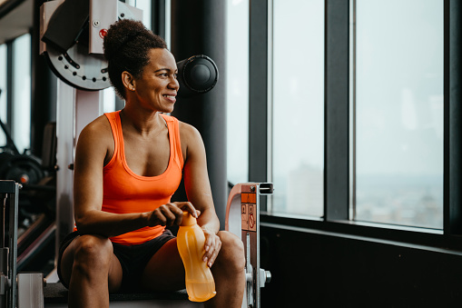 Beautiful fitness woman dressed up in sportswear sitting at gym resting after workout and thoughtfully looking into the distance while holding a water bottle.