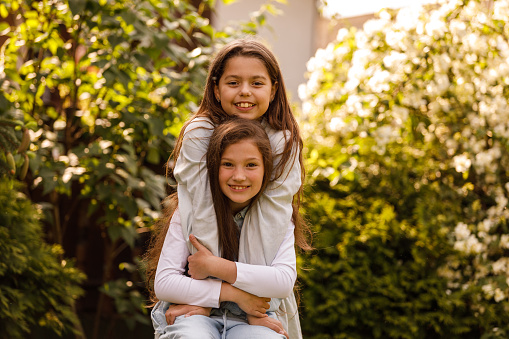 Portrait of two joyful little girls standing embraced in a sun kissed back yard with lush green foliage, looking at camera and cheerfully smiling.