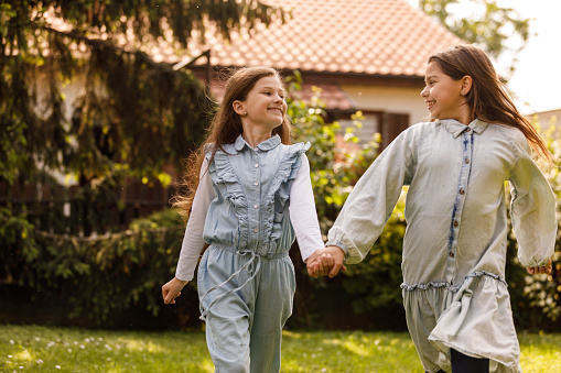 Portrait of two happy girls holding hands, walking across lush green back yard, chatting and bonding.