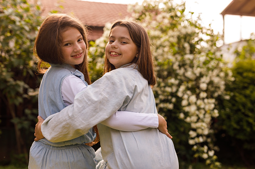 Rear view of two little girls, both with brown hair, sitting in a garden, embraced, having bonding moments. They are looking over shoulder, smiling at camera.