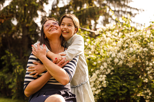 Portrait of joyful nine year old girl embracing her mother from behind while bonding, relaxing in the back yard and enjoying warm summer sunshine together.