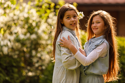 Portrait of two happy little girls standing embraced in a sunny garden, looking away, smiling and contemplating.