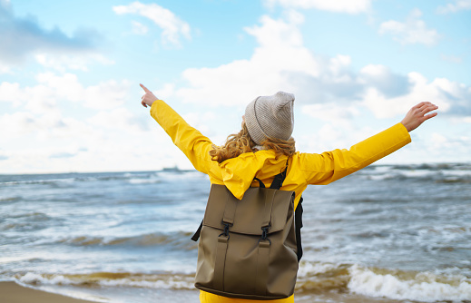 Young woman tourist in a yellow coat walks along the seashore, enjoys the seascape at sunset. Travel, tourism concept. Active lifestyle.