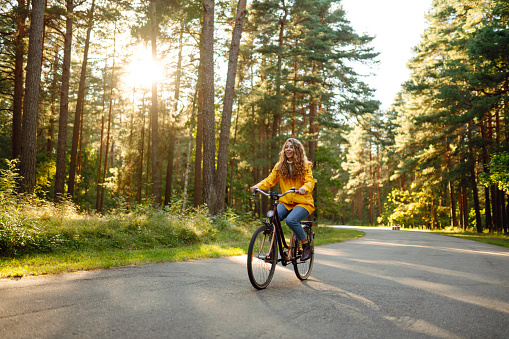 Smiling woman with curly hair in a yellow coat rides a bicycle in a sunny park. Tourist woman enjoys autumn nature. Concept of nature and active lifestyle.