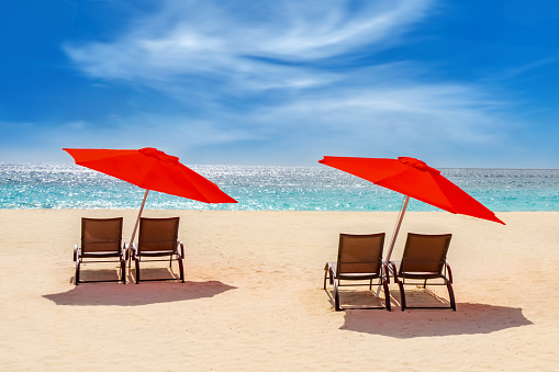 Two red beach umbrellas and reclining sunbeds on a sandy beach in the Caribbean.