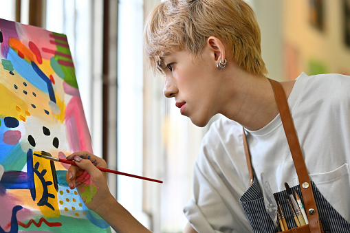 Close-up image of an Asian LGBT teenage student with colored hair doing his artwork, painting color on a canvas.