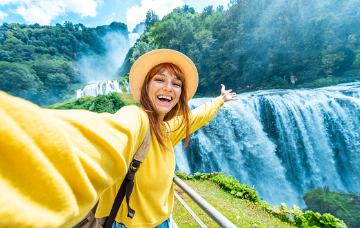 Handsome woman tourist visiting national park taking selfie picture in front of waterfall - Traveling life style concept with happy female wearing hat enjoying freedom in the nature