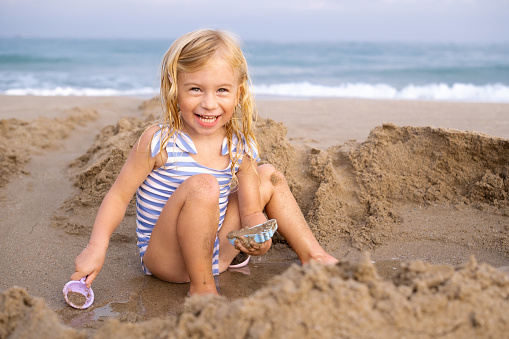 Cute laughing blonde three years old girl playing with sand at beach near sea in Spain, looking at camera.Child activity, happiness.Kid having fun at resort on vacation, holidays, tourism concept.