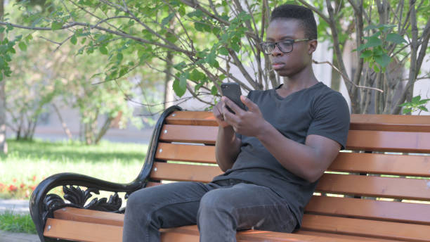 istock Young African Man using Smartphone while Sitting Outdoor on a Bench 1594391562