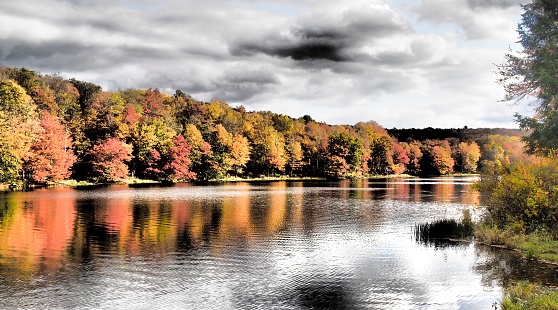 Calm lake in autumn, with dramatic cloudy sky and autumn leaf color. Reflections in water. Toned for intense mood. Waneta Lake, Sullivan County, lower Catskill Mountain Region, New York State, part of the Willowemoc Wild Forest.