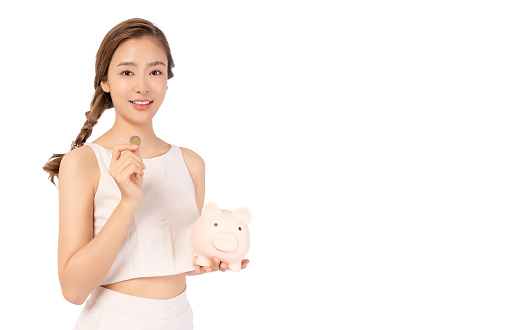 Happy young Asian woman puts coin in piggy bank for saving money She dreams become rich person in future Young girl in casual clothes holding piggy bank isolated on white background copy space