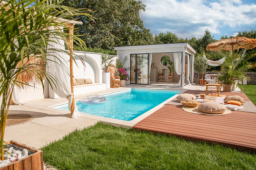 Private swimming pool area in outdoor boho garden with sun chairs on wood deck and pool house