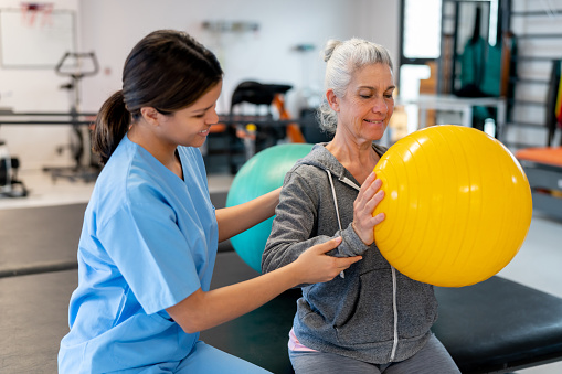 Latin American therapist correcting her patients posture during physiotherapy while patient is holding a fitness ball - healthcare concepts