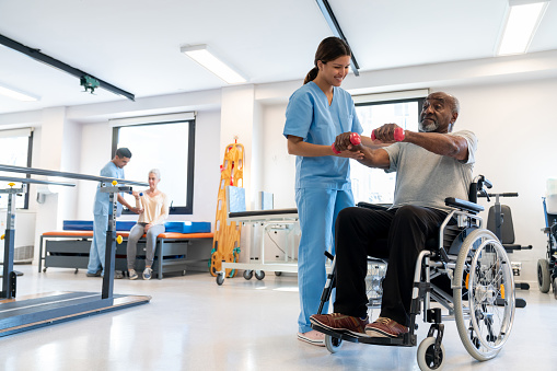 Cheerful therapist correcting the posture of her black senior patient on a wheelchair working out his arms with dumbbells - healthcare concepts - Incidental patient and therapist at background