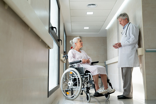 Senior doctor talking to female senior patient on a wheelchair discussing treatment while looking at her medical chart on a tablet - Healthcare concepts - Full length shot