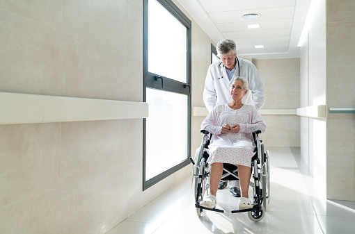 Senior doctor pushing a wheelchair with a female senior patient taking her to a procedure while talking - Healthcare concepts - Full length shot