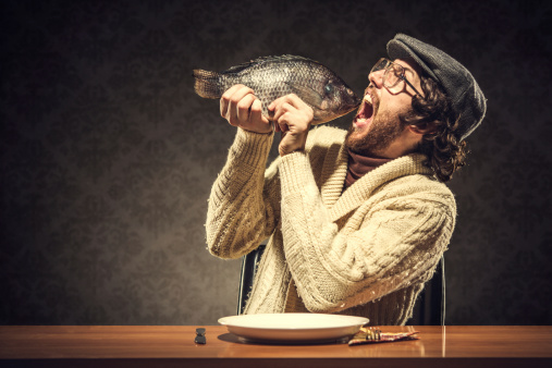 A vintage looking man wearing a newsboy hat and a turtleneck and cardigan sweater sits at the dinner table and prepares to eat a whole raw tilapia fish.  Healthy eating?  Retro damask wall paper pattern in background.  Horizontal with copy space.