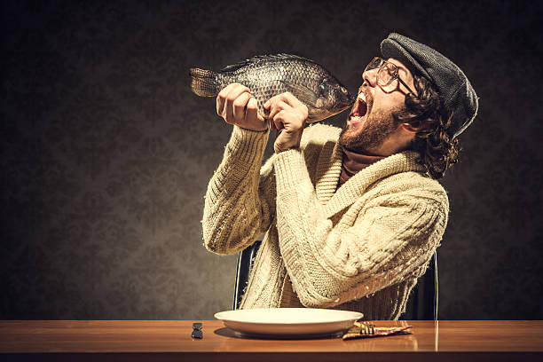 Fish Dinner A vintage looking man wearing a newsboy hat and a turtleneck and cardigan sweater sits at the dinner table and prepares to eat a whole raw tilapia fish.  Healthy eating?  Retro damask wall paper pattern in background.  Horizontal with copy space. grotesque stock pictures, royalty-free photos & images