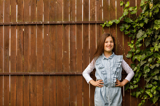 Copy space shot of happy little girl standing against a wooden fence with green foliage, hands on hips, looking at camera and smiling with confidence.