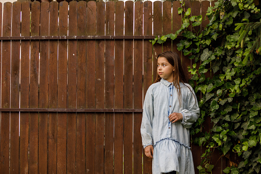 Copy space shot of happy little girl standing against a wooden fence with green foliage, looking away, smiling and daydreaming.