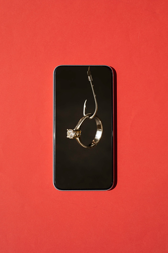 Solitaire diamond ring hanging at fishing hook in smart phone on red background. Representing online dating apps and phishing concepts.