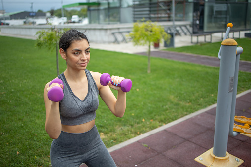 A young beautiful woman is training outside, she is doing exercises with weights in a public park