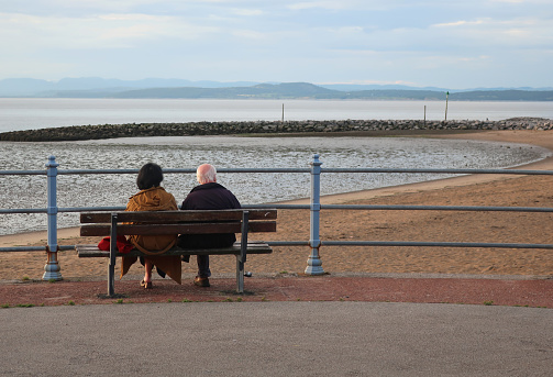 Morecambe, Lancashire, United Kingdom, July 9, 2023. Two elderly people sitting on a bench (man and woman). Looking out over Morecombe bay from the town promenade.