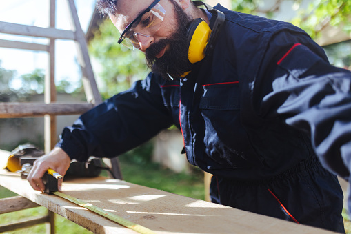 A bearded blue-collar worker measures the boards where he needs to cut them with a jigsaw