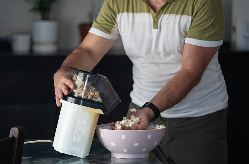 Photo of mature adult man standing in domestic kitchen and cooking popcorns with hot air popper. Shot under daylight with a full frame mirrorless camera.