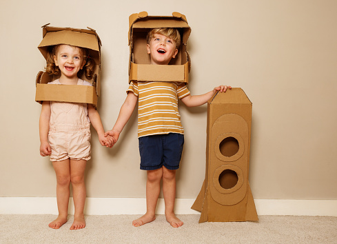 Two happy kids boy and girl stand holding hands with toy self-made rocket wearing cardboard astronaut helmets, laughing little astronauts