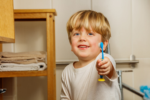 Portrait of a little blond boy brushing teeth holding toothbrush in hand and smiling to camera