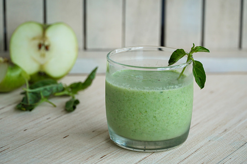 Refreshing green juice in a wooden white background, adorned with mint leaves and apple slices.