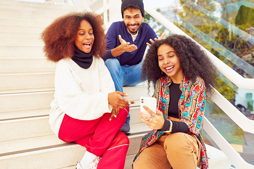 multiracial mixed group of three people young adult friends sitting on a step looking at a cell phone laughing