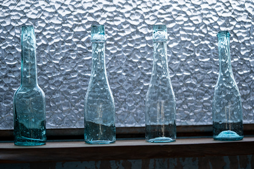 Glass bottles of shades of green, blue and amber rest on a shelf behind an outside dirty window of a Cape Cod antique shop.