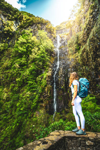istock Attractive female tourist with backpack standing on stone wall viewpoint and watches green overgrown Risco waterfall. 25 Fontes Waterfalls, Madeira Island, Portugal, Europe. 1595123775